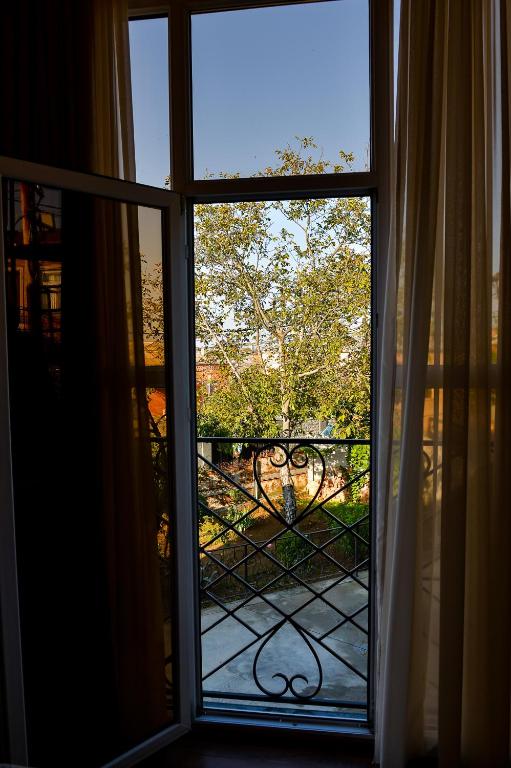 A balcony door framed by light, sheer curtains reveals a view of a lush, green tree outside. The sunlight filters through, creating a bright and airy atmosphere in the indoor space. A decorative iron railing extends outward, adding architectural interest.