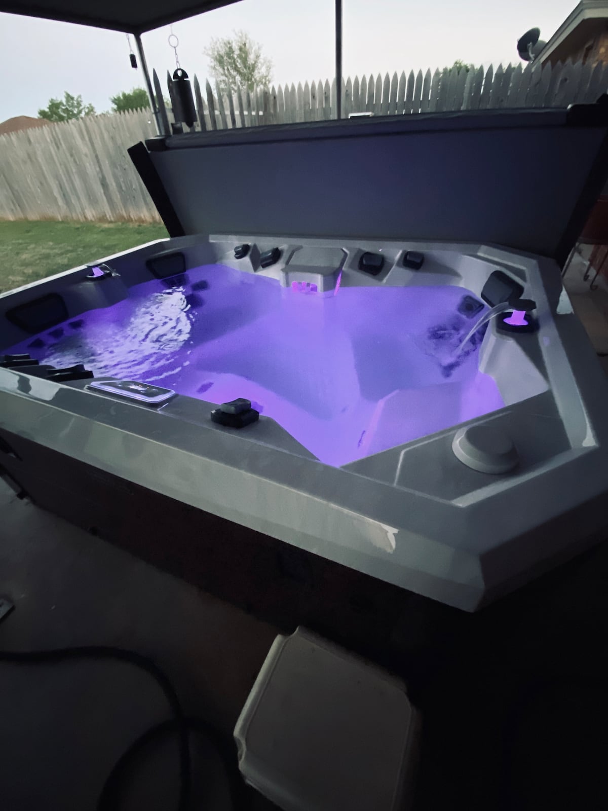 A modern hot tub is situated under a shaded patio, featuring ambient purple lighting that reflects off the water. Surrounding the tub is a spacious area with well-maintained grass visible in the background, creating a relaxing atmosphere for guests.