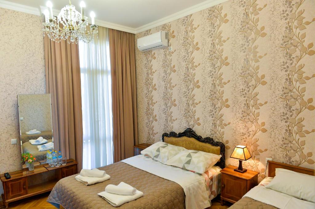 A warm-toned room features two beds adorned with neutral-colored linens. A decorative mirror reflects elegant wall patterns. A small bedside lamp adds soft lighting, while a crystal chandelier hangs above. Large drapes frame a window, allowing natural light to fill the space.