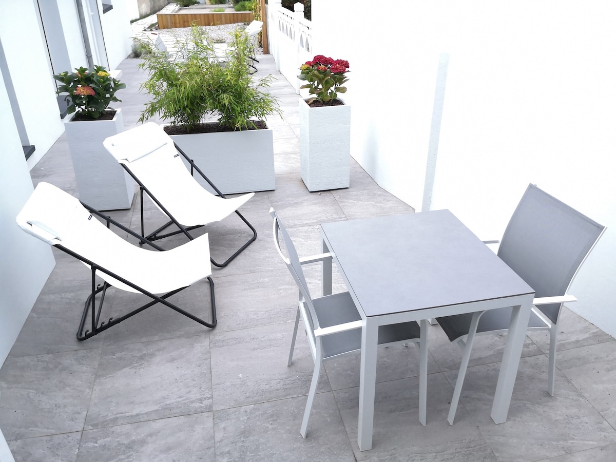 A small terrace is presented, featuring a rectangular table with four gray chairs, alongside two reclining lounge chairs. Potted plants add a touch of greenery to the light-colored tiled floor, creating a space for relaxation.
