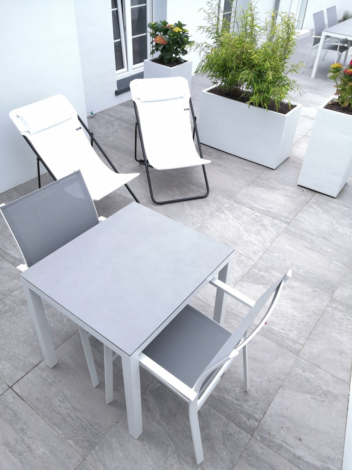 A small terrace is presented, featuring a gray table and a matching chair. Two reclining chairs are positioned nearby, with green plants in white planters enhancing the serene atmosphere. The smooth stone tiles complete the outdoor space.