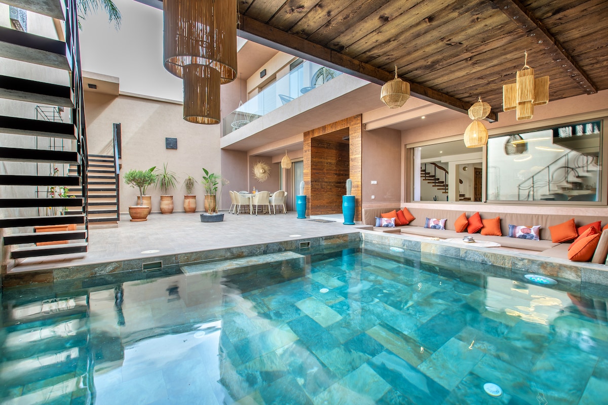 An inviting indoor space features a sparkling pool surrounded by elegant decor. Natural wood beams accent the ceiling, while soft lighting from hanging fixtures creates a tranquil atmosphere. A comfortable seating area with colorful cushions offers a cozy spot for relaxation, with views of the open living area.