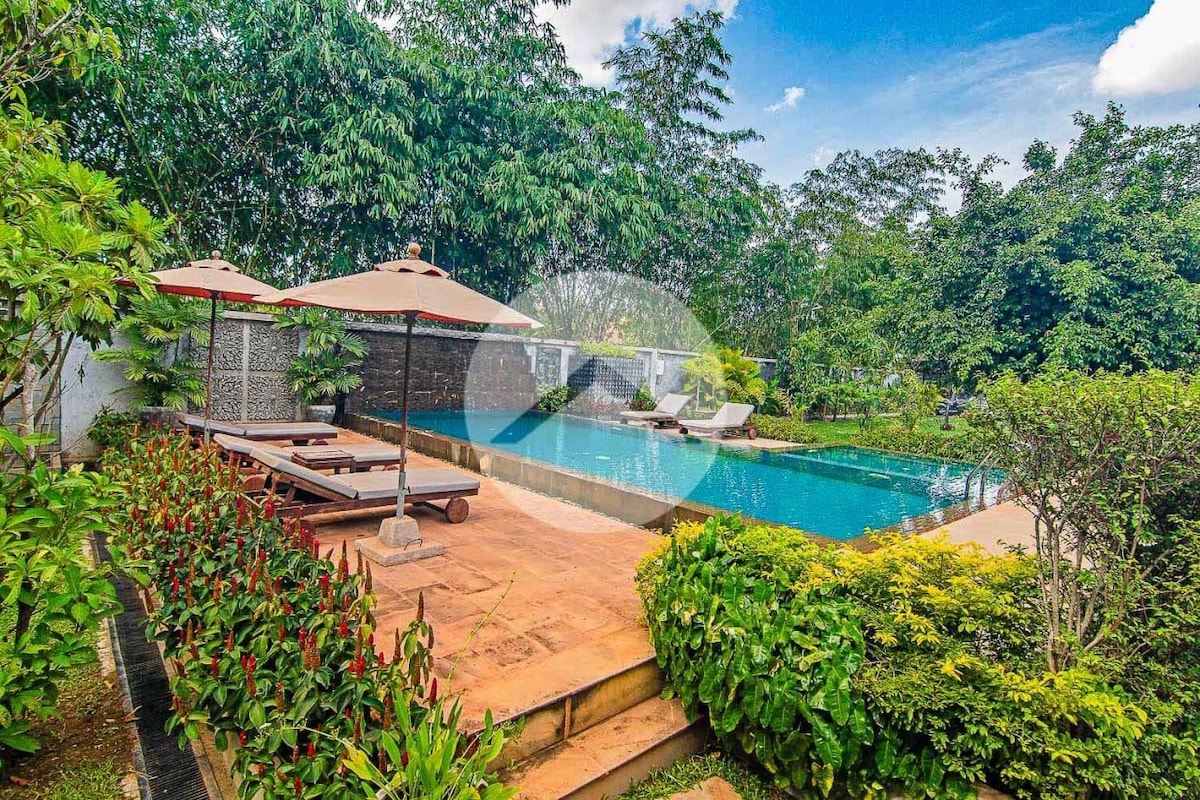 A private pool is surrounded by lush greenery, providing a serene outdoor escape. Sun loungers are positioned under shaded umbrellas, and vibrant plants line the pathway leading to the water's edge, creating a harmonious blend of relaxation and nature.