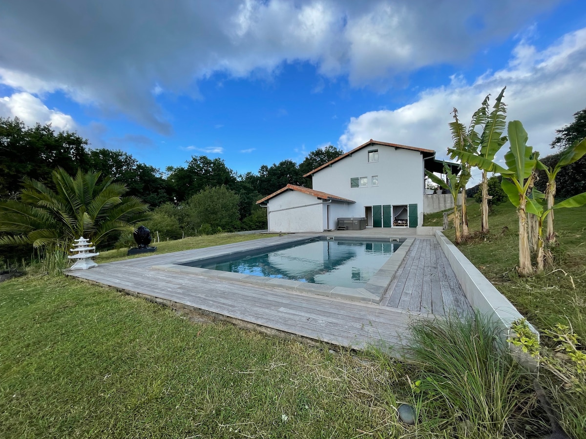 A modern villa is set amidst lush greenery, featuring a rectangular pool surrounded by a wooden deck. The building's white facade is complemented by a tiled roof, while tropical plants frame the scene, offering a serene view of the natural surroundings.