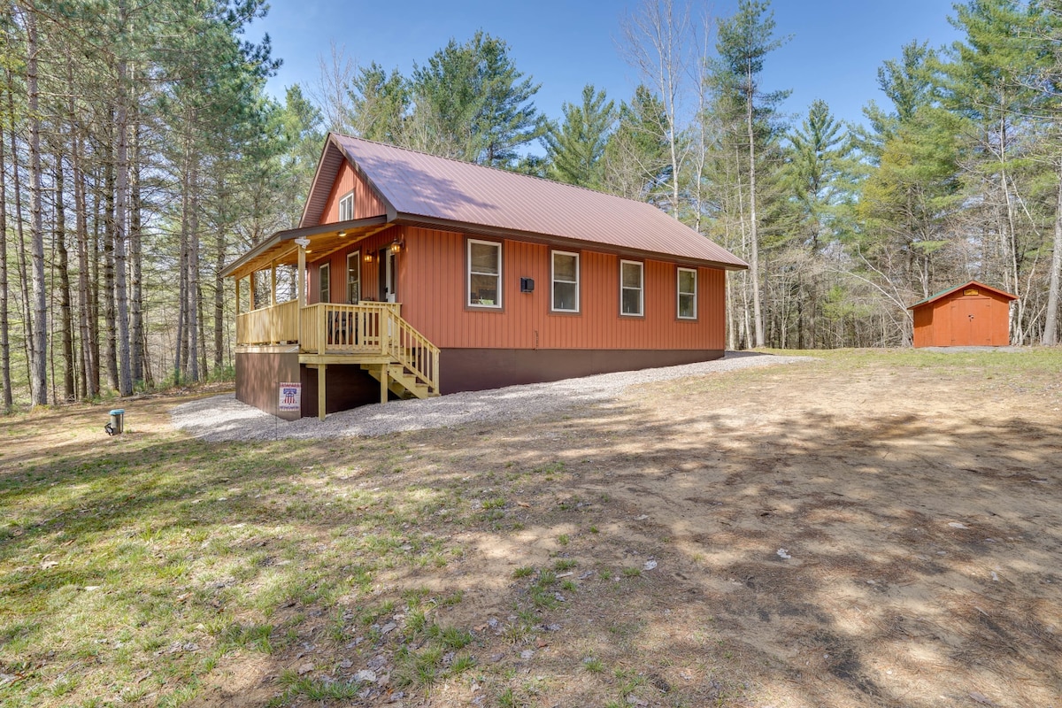The exterior view of the vacation rental showcases a charming orange house with a metal roof, surrounded by tall trees. A wooden porch with steps invites entry, while a small storage shed is visible in the background on the 1.5-acre lot.
