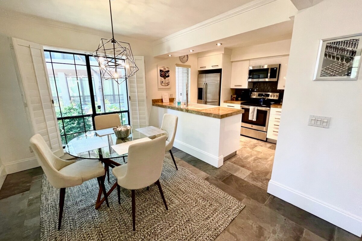 A modern dining area features a round glass table surrounded by four upholstered chairs. Natural light filters through large windows, highlighting the sleek kitchen behind, equipped with stainless steel appliances and a contemporary design. A textured rug covers the floor, adding warmth to the space.