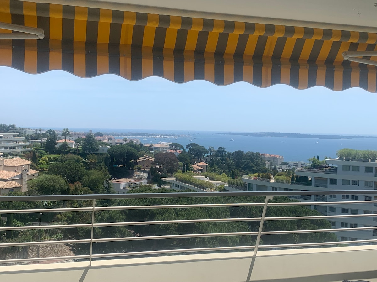 A panoramic sea view is framed by a striped awning, highlighting the coastline dotted with yachts. Lush greenery and residential areas are visible in the foreground, creating a serene backdrop against the shimmering water.