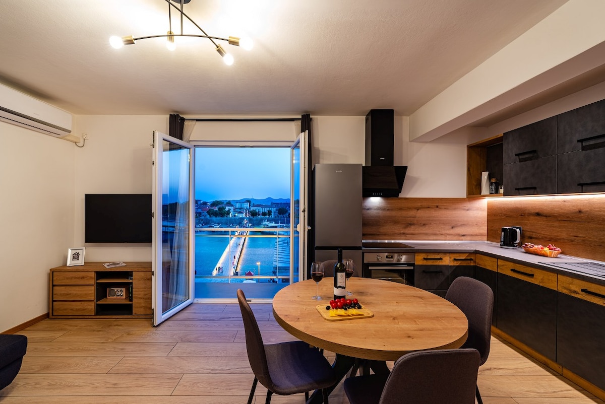 An open-plan kitchen and dining area features a round wooden table surrounded by four grey chairs. Large glass doors open to a view of the waterfront, with city lights beginning to illuminate the scene at dusk. A modern kitchen with sleek black cabinets is also visible.