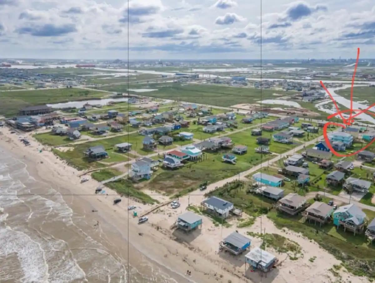 An aerial view captures a coastal neighborhood, showcasing a mix of residential homes and green spaces. The Gulf of Mexico is visible at the bottom, with sandy shores lined by various structures, indicating proximity to the beach. The highlighted unit is seen among the neighborhood.