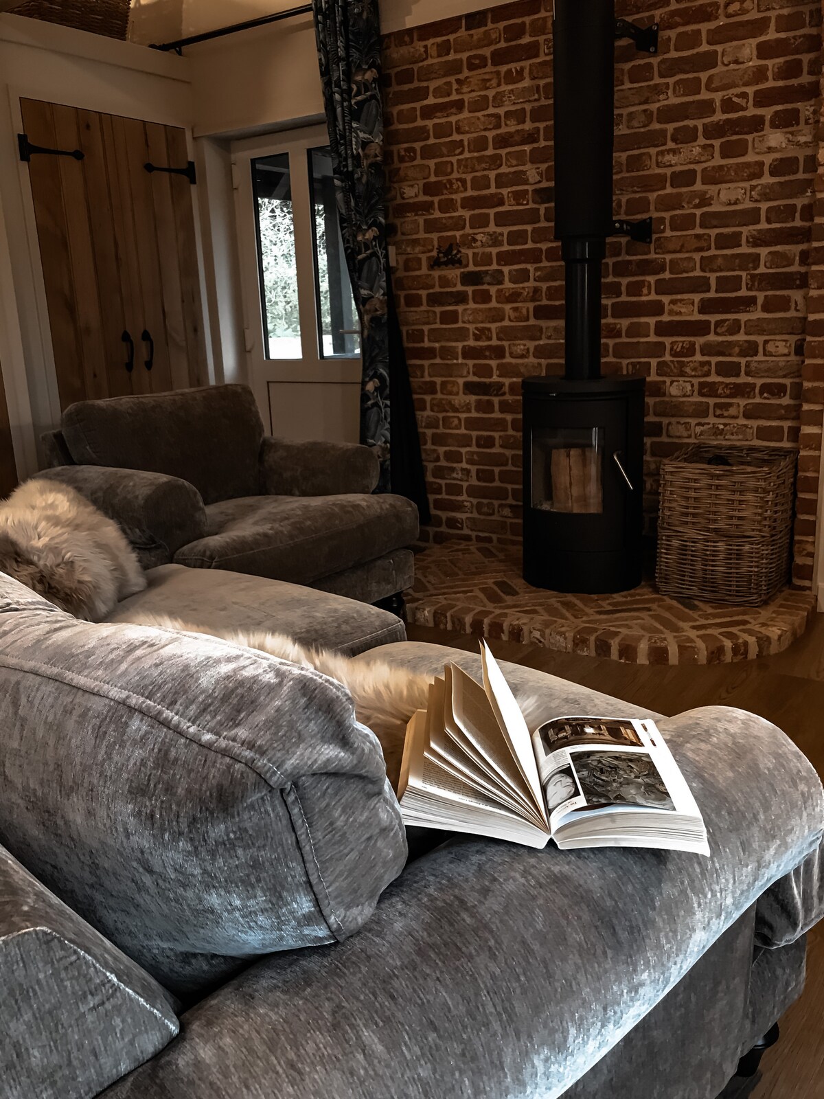 A cozy living area features a plush, grey sofa beside a rustic brick wall. A black wood-burning stove stands in the corner, with a woven basket nearby. An open book rests on the sofa, inviting guests to relax and unwind.