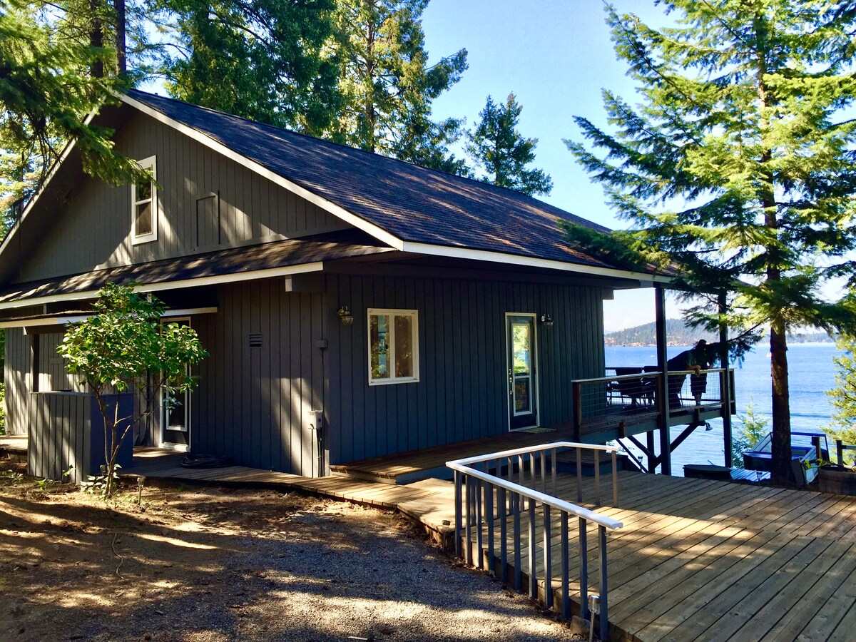 A modern, two-story house sits among tall pine trees, featuring a slightly sloped roof and large windows. A wooden deck extends from the side, providing views of the nearby lake. The property showcases a serene waterfront setting, ideal for relaxation.