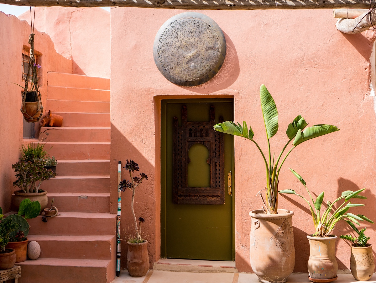 A traditional entrance is framed by a green door and adorned with intricate artwork. Several potted plants in clay pots enhance the setting. Stairs ascend to the left, leading to additional spaces within the camp, with warm earthy tones on the walls creating a welcoming atmosphere.