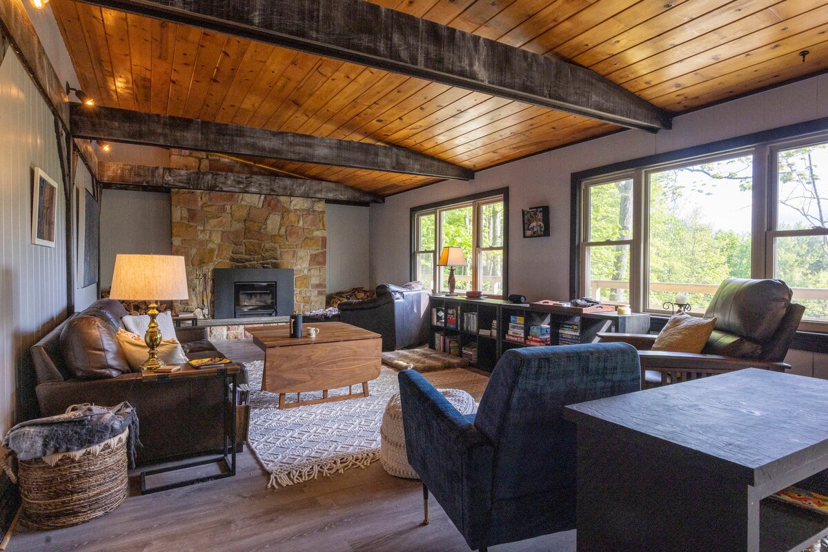 A comfortable living room features a stone fireplace as the focal point. Large windows provide natural light and views of the surrounding greenery. A mixture of seating options, including two armchairs and a cozy sofa, are arranged around a wooden coffee table.