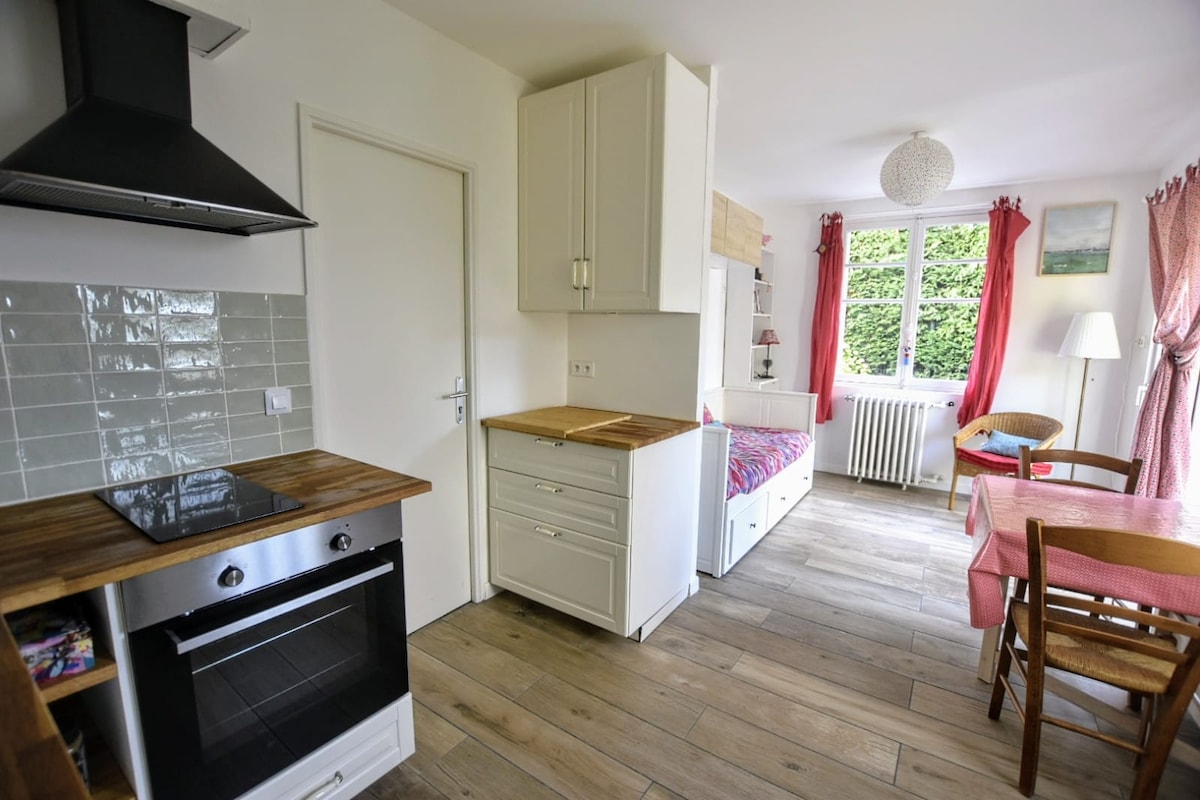 A well-equipped kitchen area is visible, featuring a stovetop and countertop with wooden accents. A cozy sleeping area is situated to the right, with a bed adorned in vibrant bedding. Natural light fills the space through the window, complemented by light curtains and a soft glow from a nearby lamp.