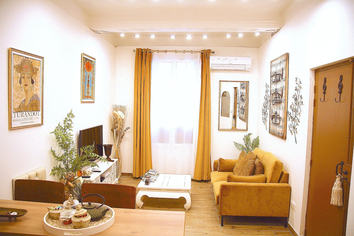 The living area features a warm color scheme with yellow curtains framing a large window. A comfortable mustard-colored sofa and a light-colored coffee table are present, complemented by decorative wall art. A TV is positioned on a white media console, and potted plants add a touch of greenery.
