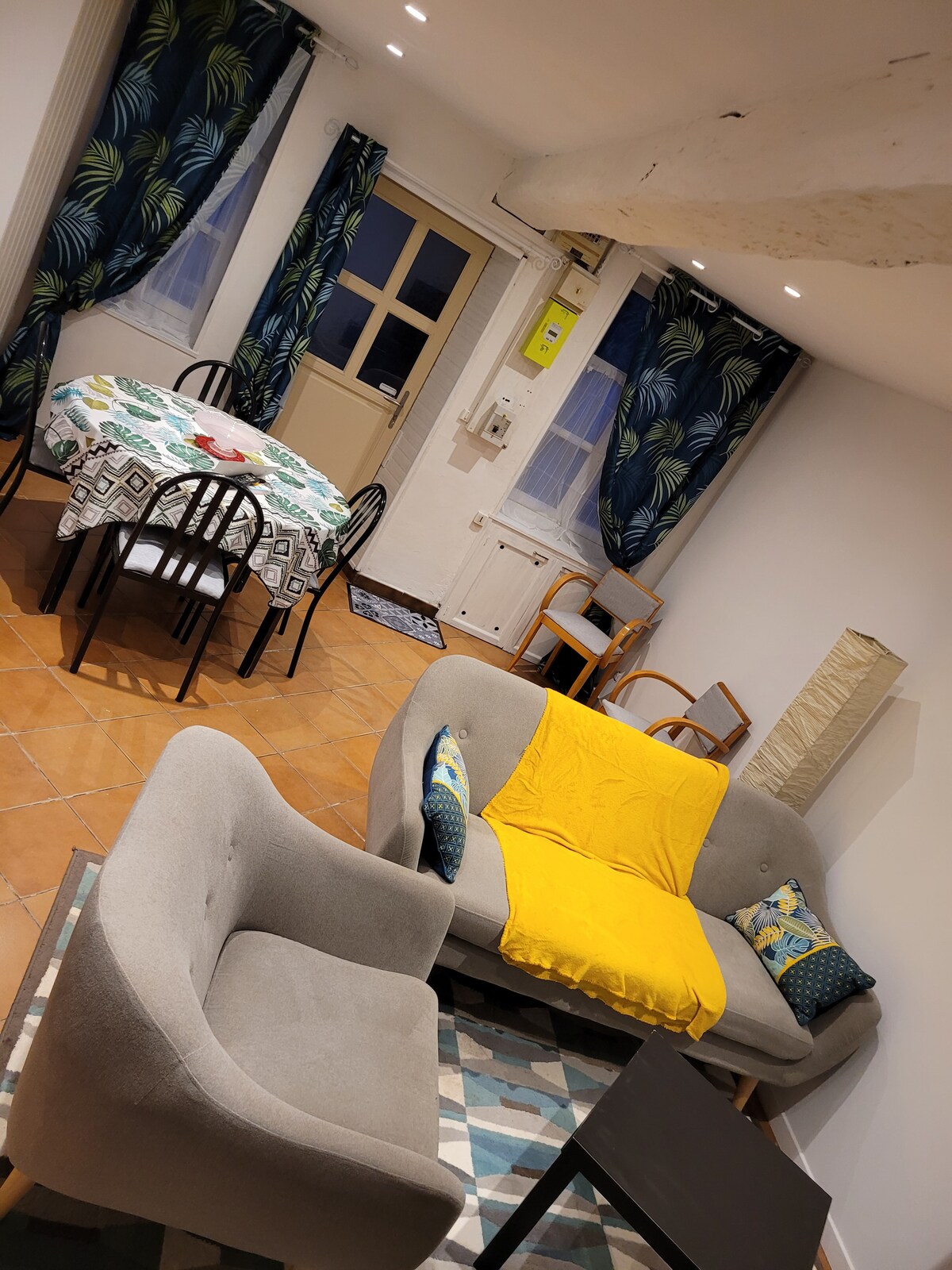 A cozy living area displays a grey sofa adorned with a yellow throw and patterned cushions. A round dining table is set with patterned tableware, surrounded by black chairs. Bright blue curtains frame the window, while light illuminates the terracotta tiled floor.
