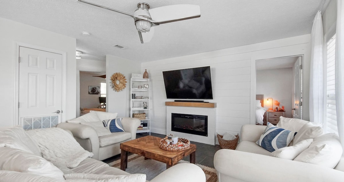 A light-filled living area features a cozy arrangement of soft sofas and a wooden coffee table. A mounted television hangs above a modern electric fireplace. Large windows allow natural light to brighten the space, while tasteful decor adds a relaxed feel.