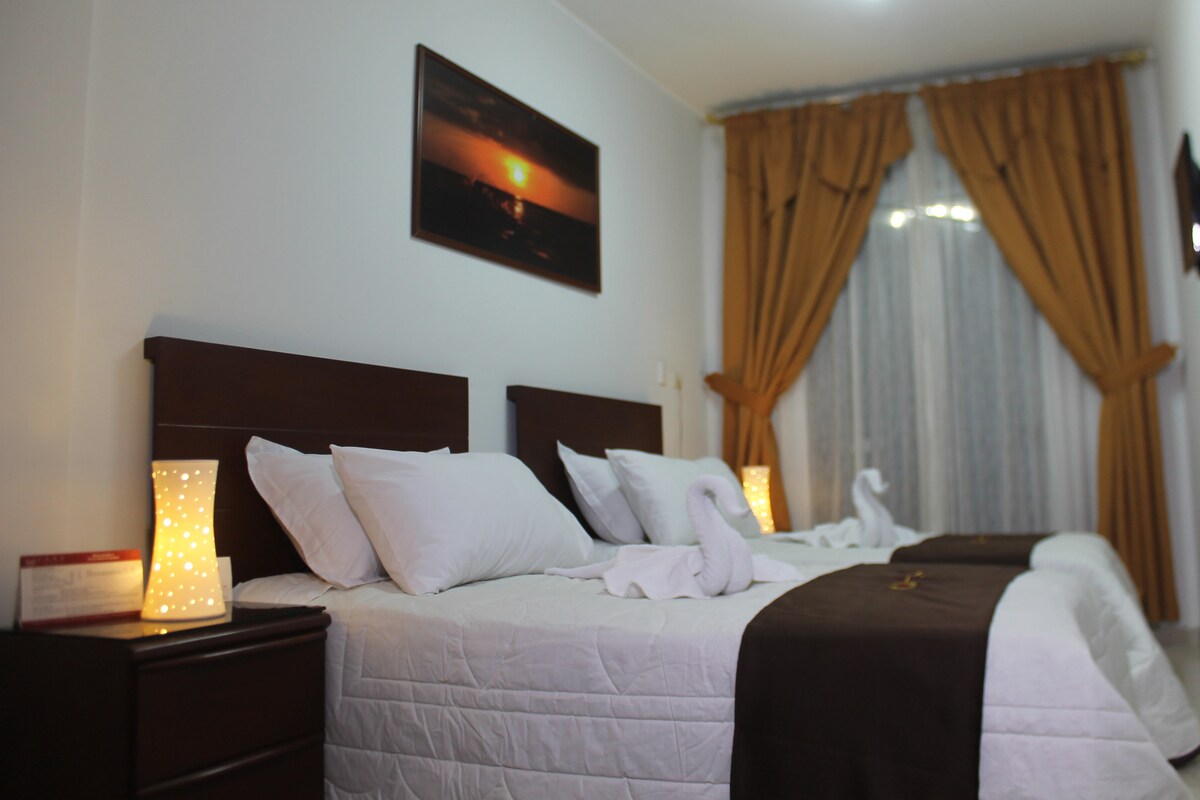 A spacious sleeping area features two neatly made beds with plush white linens. Soft lighting is provided by decorative lamps on either side, and warm curtains frame a large window, allowing natural light to enhance the inviting ambiance.