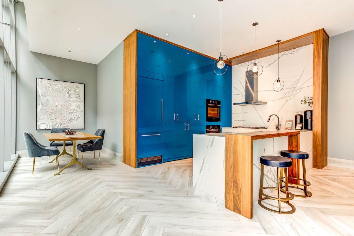 The kitchen area features modern blue cabinetry paired with a marble countertop and backsplash. Two bar stools are positioned at the kitchen island, while a small dining table with stylish chairs is located nearby. Natural light fills the space through large windows.