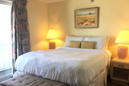 A bright and inviting room features a spacious bed with white linens and decorative pillows. Flanking the bed are two bedside lamps casting a warm glow. A soft bench is positioned at the foot of the bed, and a framed artwork adorns the wall above.