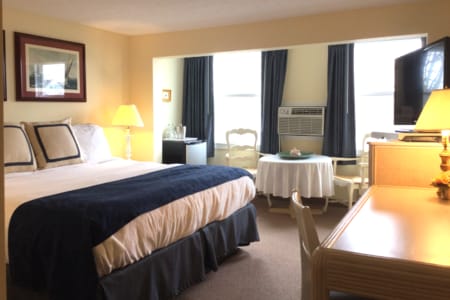 A spacious suite features a king sized bed dressed in white linens with a navy blanket. A seating area with a small table is positioned near the window. An air conditioning unit is visible, and a flat screen TV is mounted on the wall.