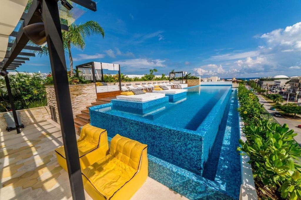 An elegant rooftop pool area features a serene infinity pool with blue mosaic tiles. Yellow loungers are positioned around the poolside, providing a relaxing space. Lush green landscaping surrounds the area, enhancing the inviting atmosphere against a clear blue sky.