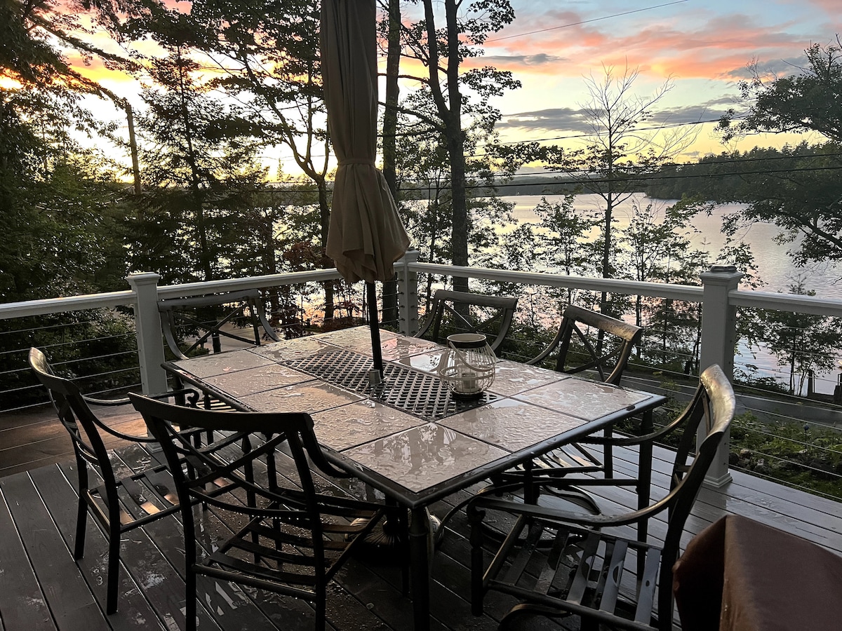 An outdoor dining set is positioned on a spacious deck, featuring a large table surrounded by six chairs. The backdrop includes tranquil lake views, with trees framing the scene. A soft, warm light glows from a lantern on the table, complemented by a setting sun.