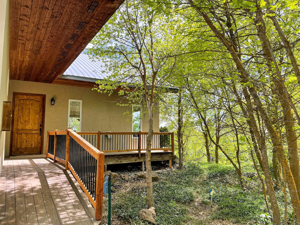A wooden deck extends from the entrance, surrounded by lush greenery and trees. Natural light filters through the foliage, creating a peaceful outdoor setting. The entrance door, crafted from wood, features a rustic design, complementing the serene environment.