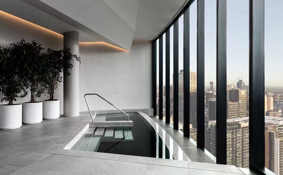 An indoor spa pool is situated beside floor-to-ceiling windows, offering expansive views of the city skyline. The space features sleek tile flooring and modern design elements, with large potted plants adding a touch of greenery to the serene environment.