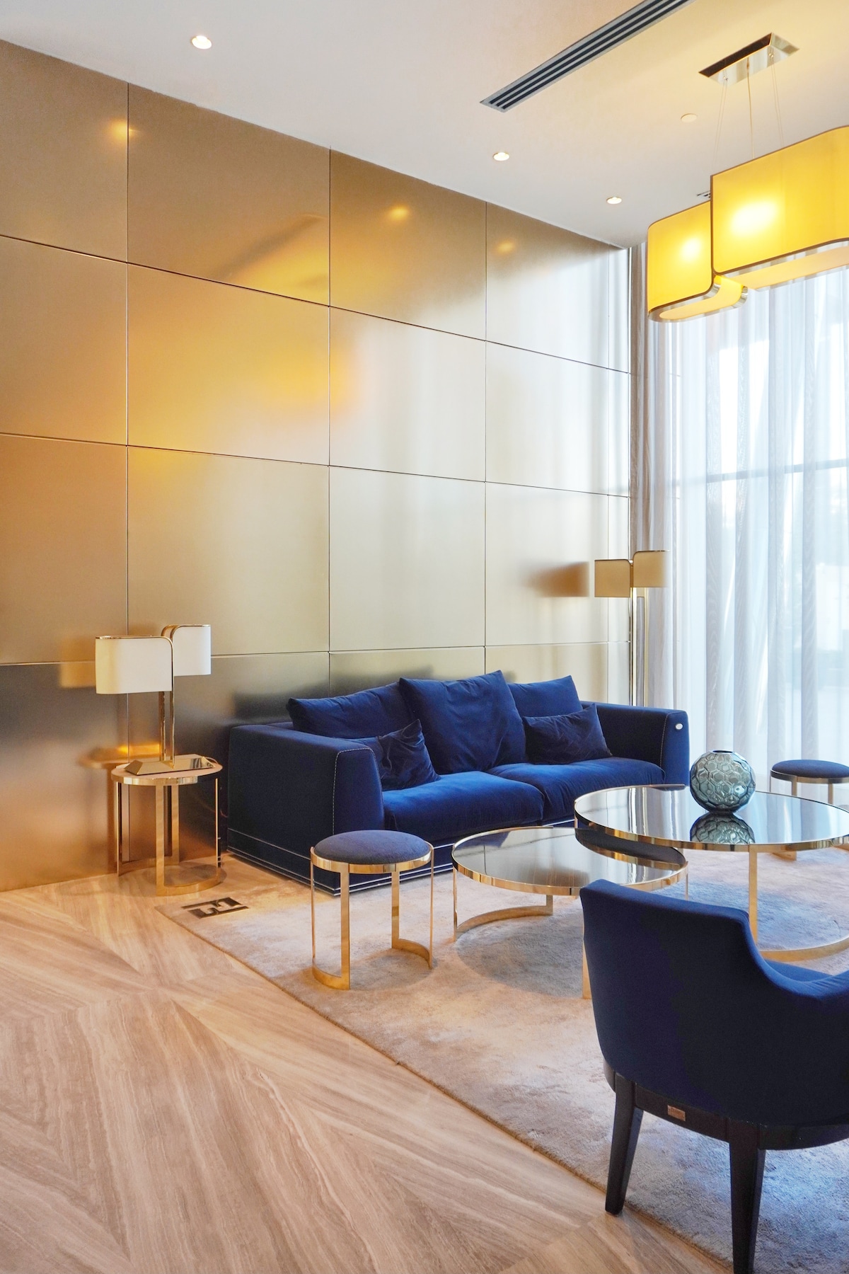 A modern living area is highlighted by gold-toned wall panels and soft lighting. A blue sofa is complemented by glass-top tables and sleek chairs. Natural light streams through large windows, enhancing the spacious feel of the room.