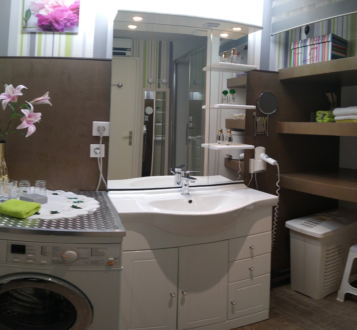 The well-appointed bathroom features a modern sink unit with storage beneath and a washing machine integrated into the design. A wall-mounted mirror and shelves are seen, holding toiletries and decorative items. A towel rack and floral accents bring a touch of style to the space.