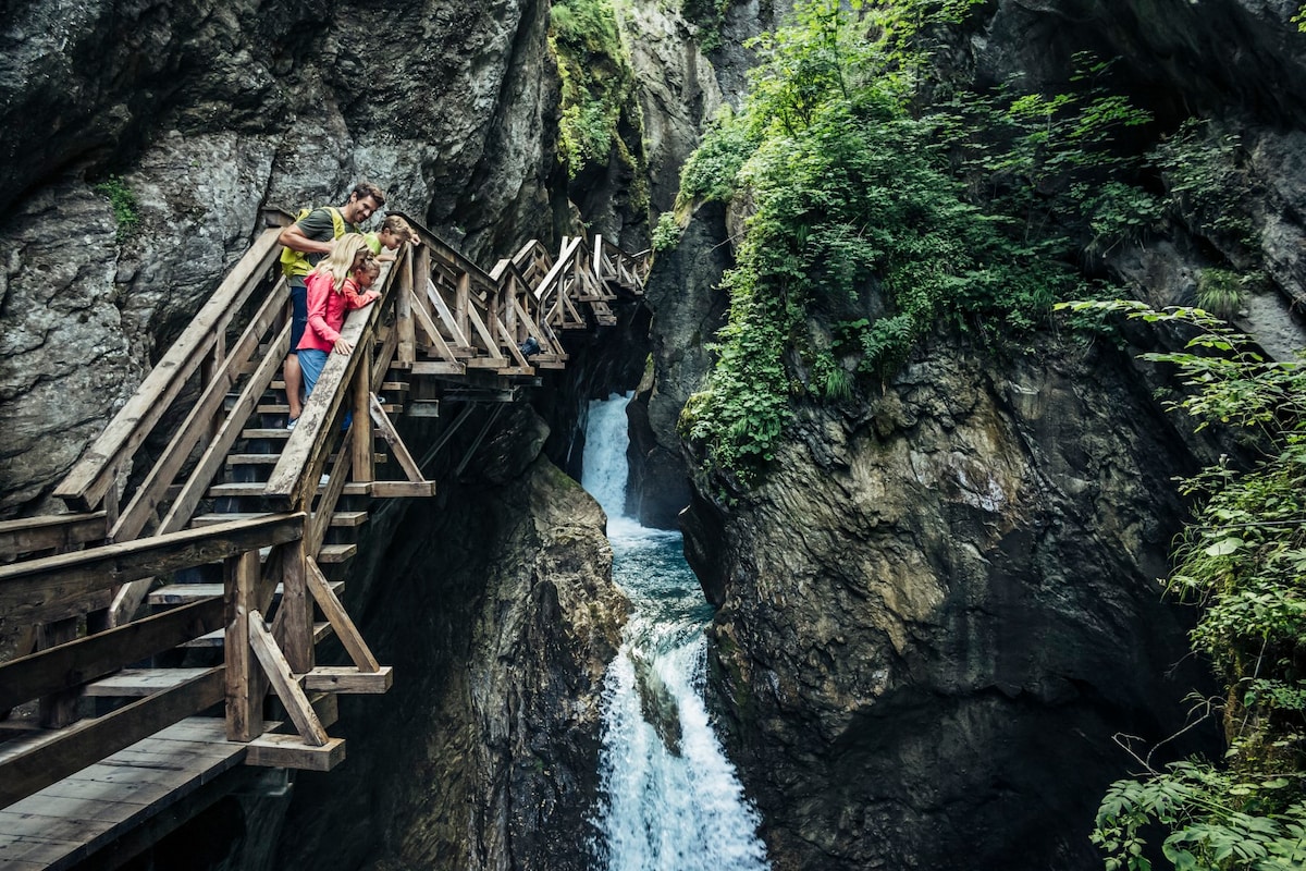 A wooden walkway winds through a rocky gorge, flanked by steep cliffs and lush greenery. Water cascades down from a hidden waterfall below, creating a serene focal point in the natural landscape. The structure allows for safe passage while providing scenic views.