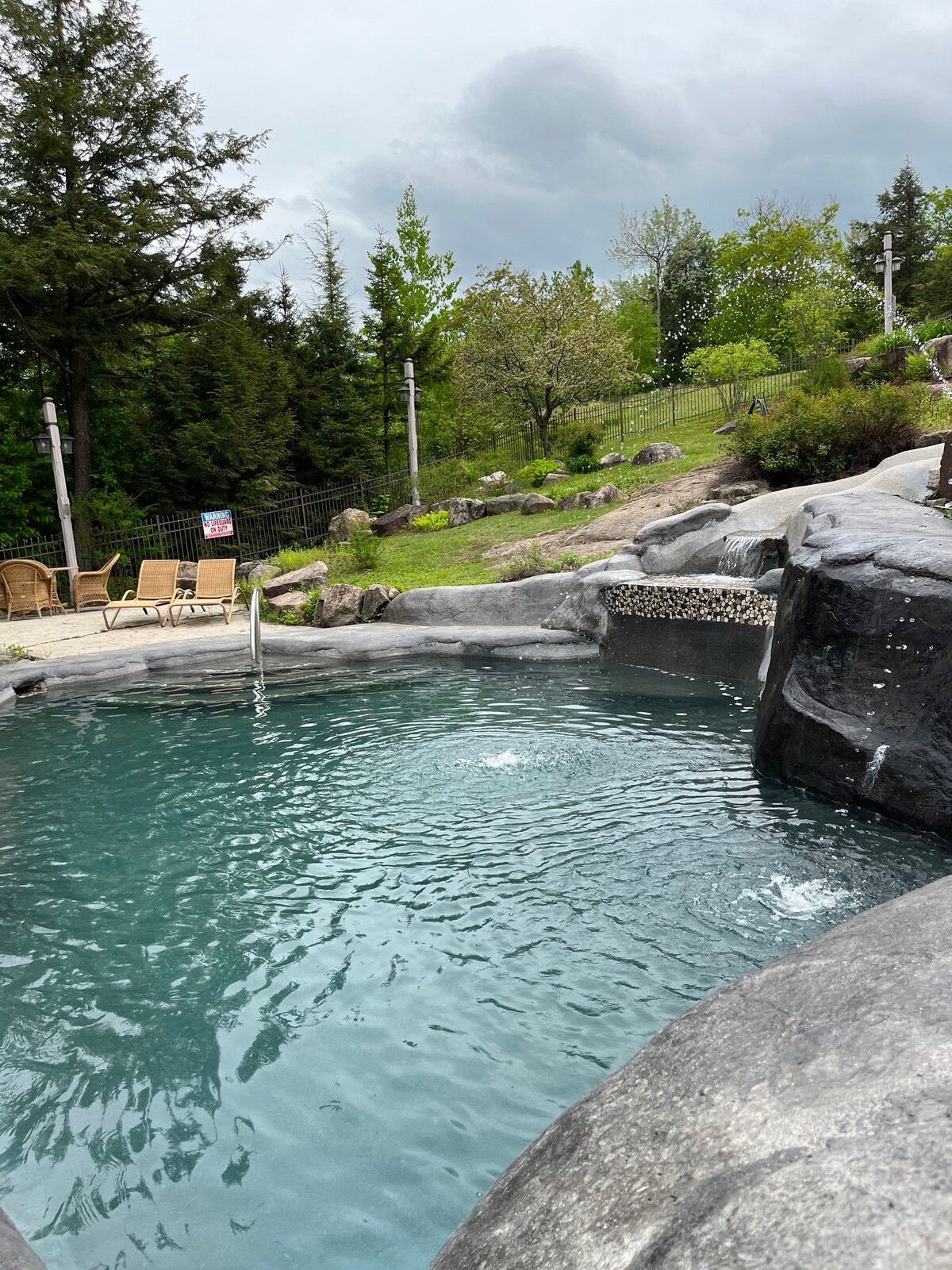 A natural rock formation surrounds a shallow pool, with water creating gentle ripples. Comfortable lounge chairs are positioned nearby, nestled among greenery. Trees and shrubs provide a serene backdrop, enhancing the ambiance of a tranquil outdoor space.