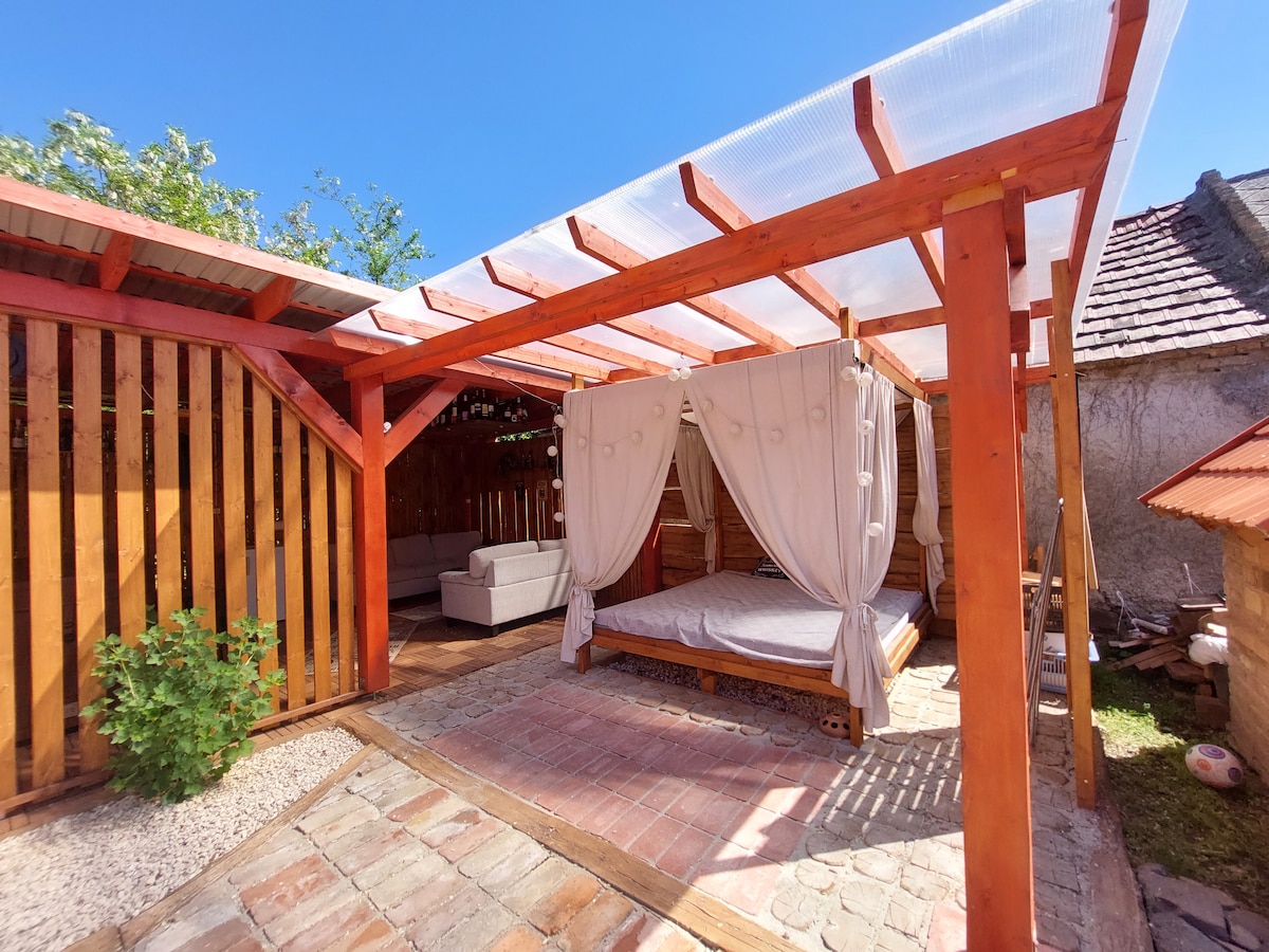 An outdoor area features a spacious bed with soft drapes, inviting relaxation. The wooden structure is complemented by a comfortable seating area and surrounded by gravel and greenery, creating a serene space for rest and leisure.