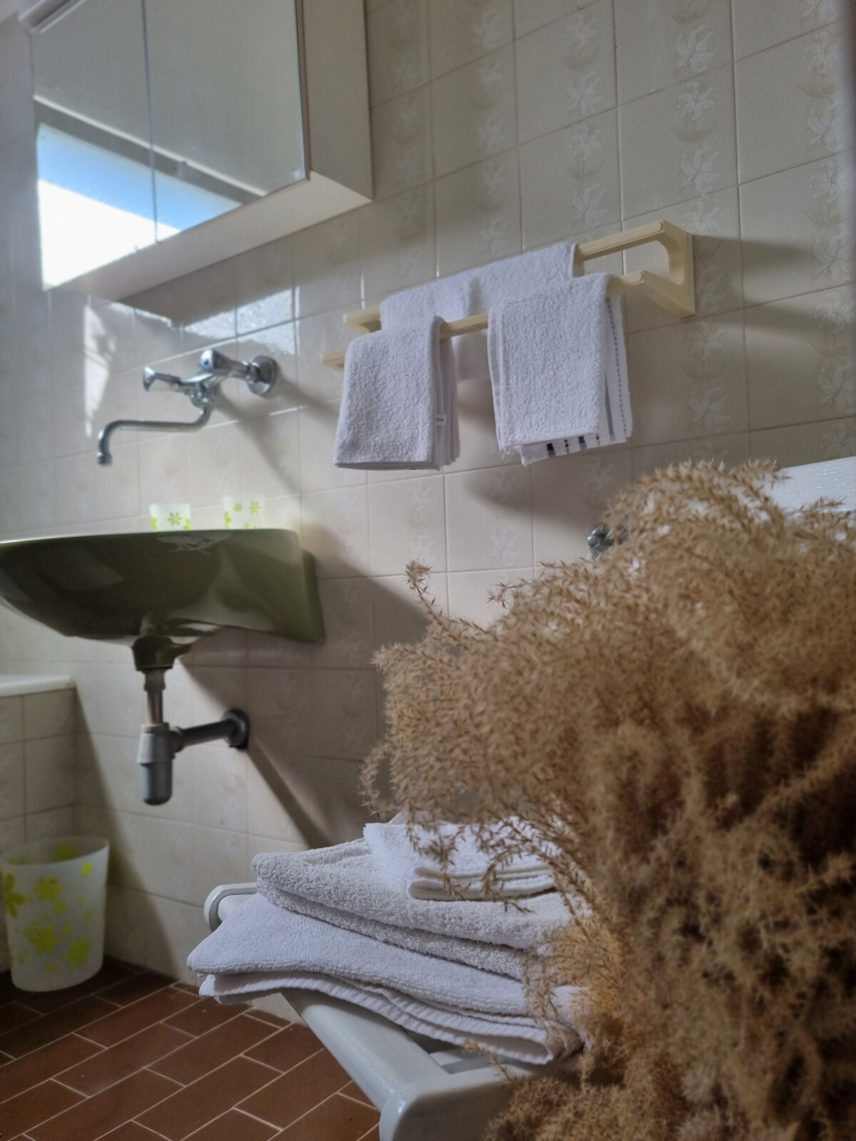 A private bathroom is presented, featuring a green sink and a wall-mounted mirror. White towels are neatly hung on a rack, while a small white bin with a floral design is placed nearby. Fresh towels are stacked on a shelf, accompanied by a bouquet of dried flowers.