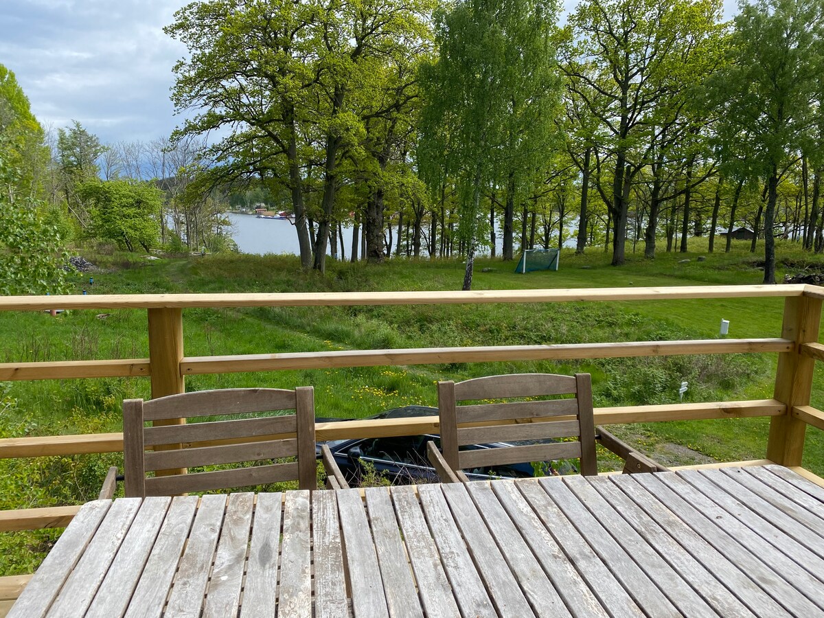 Outdoor seating is arranged on a wooden deck, featuring a rectangular table and several chairs. A view of lush greenery and the water is visible in the background, framed by trees. Sunlight filters through the foliage, creating a serene atmosphere.