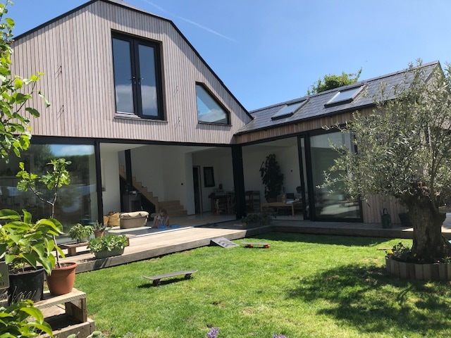 The exterior showcases a modern two-story home with a combination of wood and glass elements. Large windows allow natural light to fill the interior spaces. A well-maintained grassy area is surrounded by potted plants and features a small patio for outdoor enjoyment.