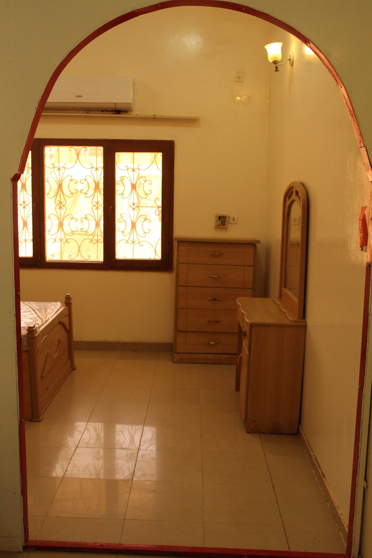 A bedroom entrance is framed by an arched doorway. Natural light filters through a set of decorative windows, illuminating the wooden bed and matching dresser. A mirror is positioned on another dresser, enhancing the sense of space within the room.