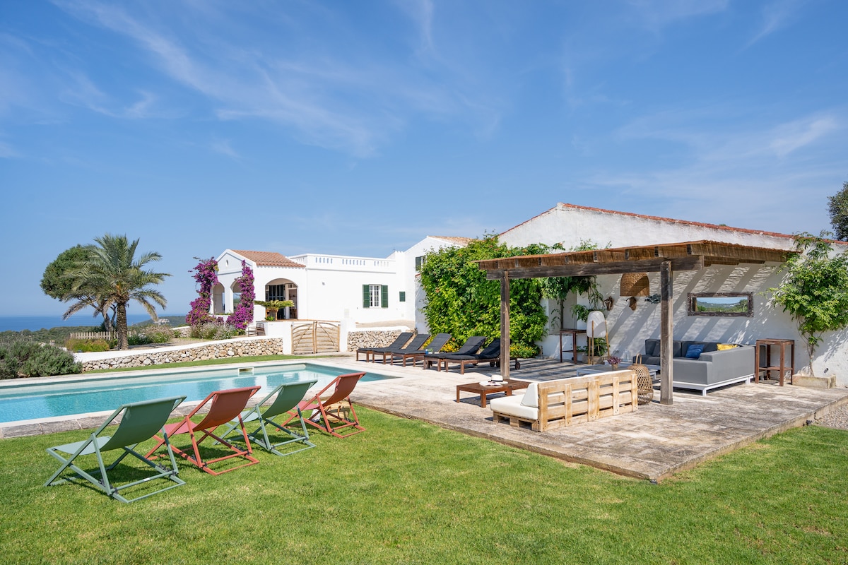 An outdoor area is presented with a spacious pool surrounded by sun loungers. A shaded pergola offers seating options for relaxation. Lush greenery and blooming plants accent the setting, while the house in the background features white walls and a traditional design, complemented by palm trees.