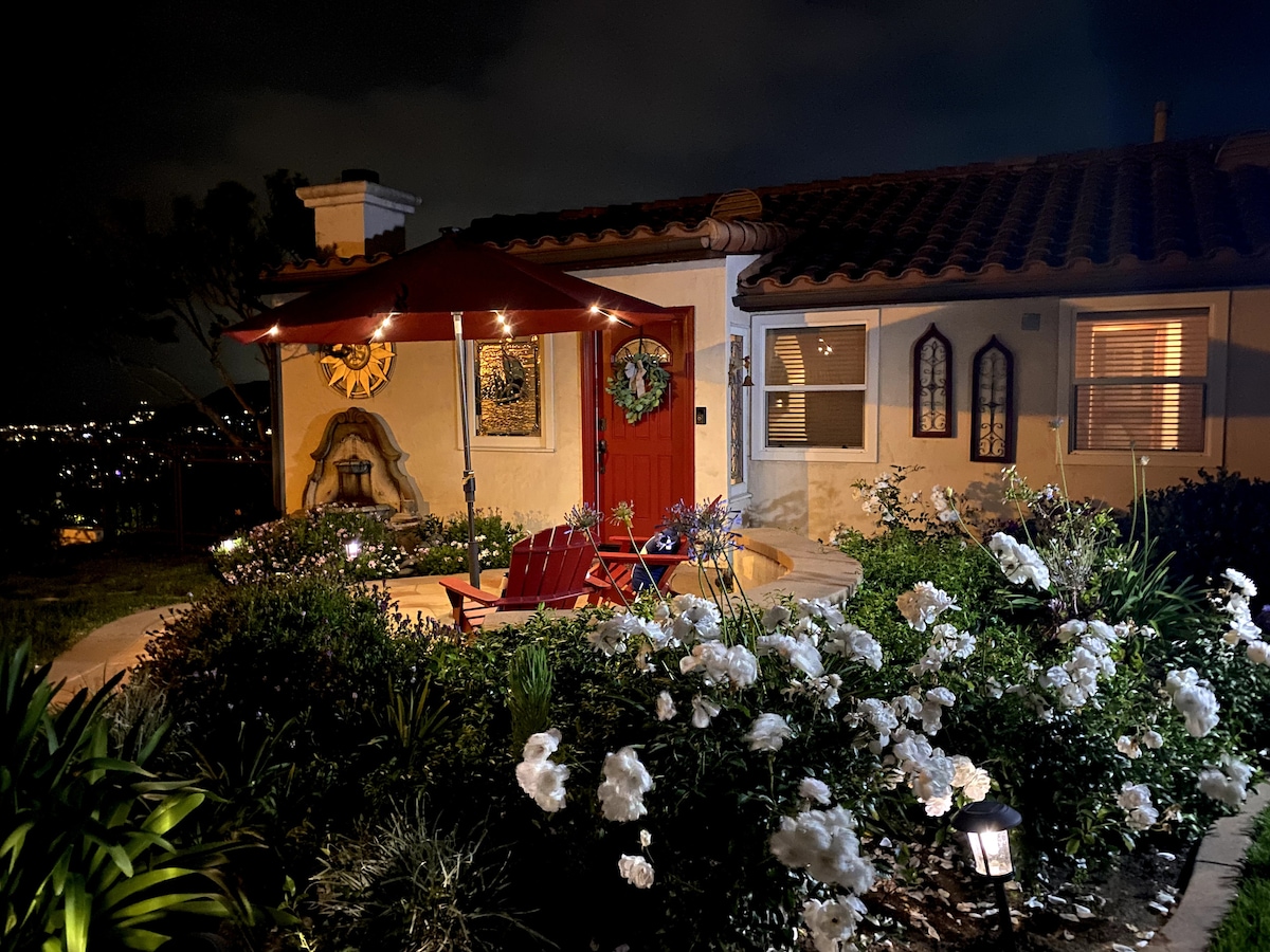 A cozy outdoor seating area is illuminated by soft lights beneath a red umbrella. Surrounding the space, white roses bloom alongside shrubs, enhancing the serene ambiance. The entrance features a terracotta tile roof and distinctive arched windows, contributing to the charming, Spanish-style architecture.