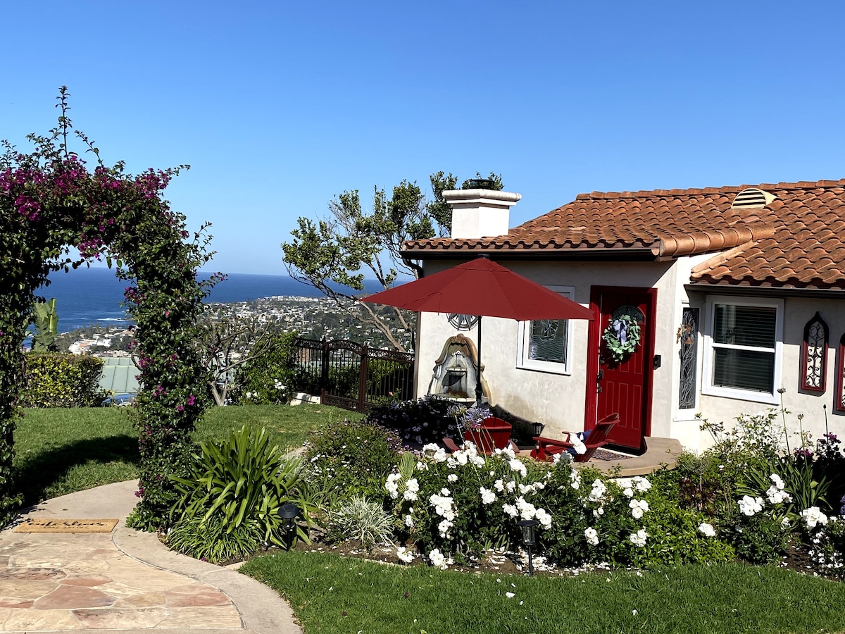 The entrance of the house is framed by blooming bougainvillea and white flowers, leading to a welcoming red door. A patio area is shaded by a wide umbrella with two chairs positioned nearby, providing a view of the ocean beyond.