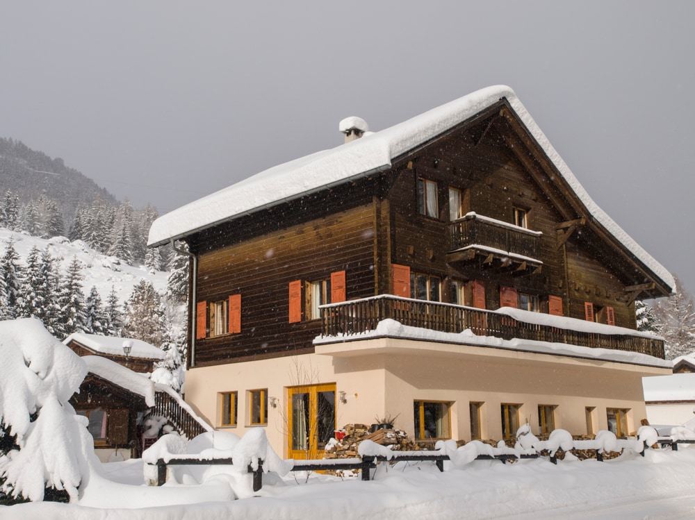 The chalet features a traditional wooden facade, accented by a balcony and orange shutters. A thick blanket of snow covers the roof and surrounding landscape, creating a serene winter scene. Towering trees are visible in the background, adding to the picturesque ambiance.