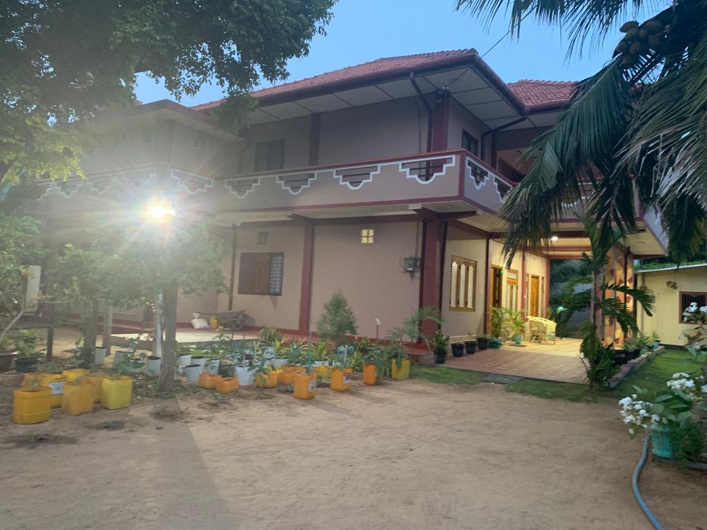 A spacious home is set amidst a well-maintained garden featuring various potted plants. The exterior showcases a traditional architectural style with a red-tiled roof and wooden accents. A clear path leads to the entrance, illuminated by soft lighting in the evening.