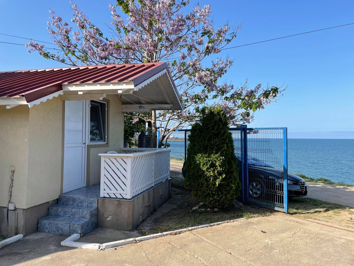 The exterior of the bungalow is shown, featuring a small covered porch with a railing. A flowering tree is visible nearby, and a clear view of the sea extends beyond the driveway, where a car is parked. The setting conveys a relaxed atmosphere.