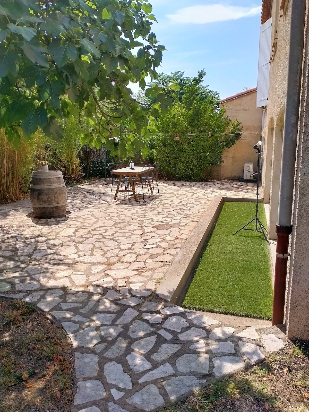 A private outdoor space features a stone-paved patio surrounded by greenery. A wooden table with chairs is positioned near a decorative barrel. A narrow strip of artificial grass adds to the setting, with sunlight filtering through tree branches overhead.