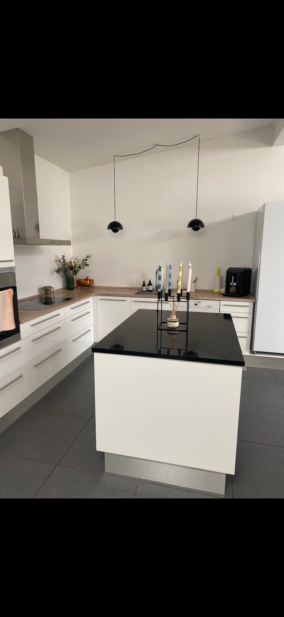 A modern kitchen is presented with sleek white cabinetry and a dark stone countertop. An island with seating is centered in the space, featuring a candelabra with five colored candles. Light fixtures hang above, and appliances are arranged along the walls.