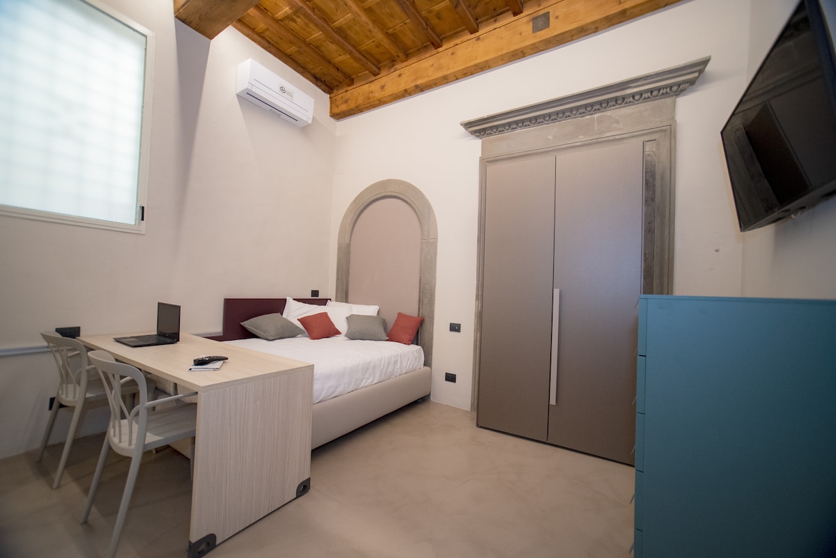 A renewed studio is displayed, featuring a spacious bed with soft linens and decorative pillows. A compact table and two chairs are situated nearby, along with a laptop on the table. An air conditioning unit is mounted on the wall, and a flat-screen TV is visible.