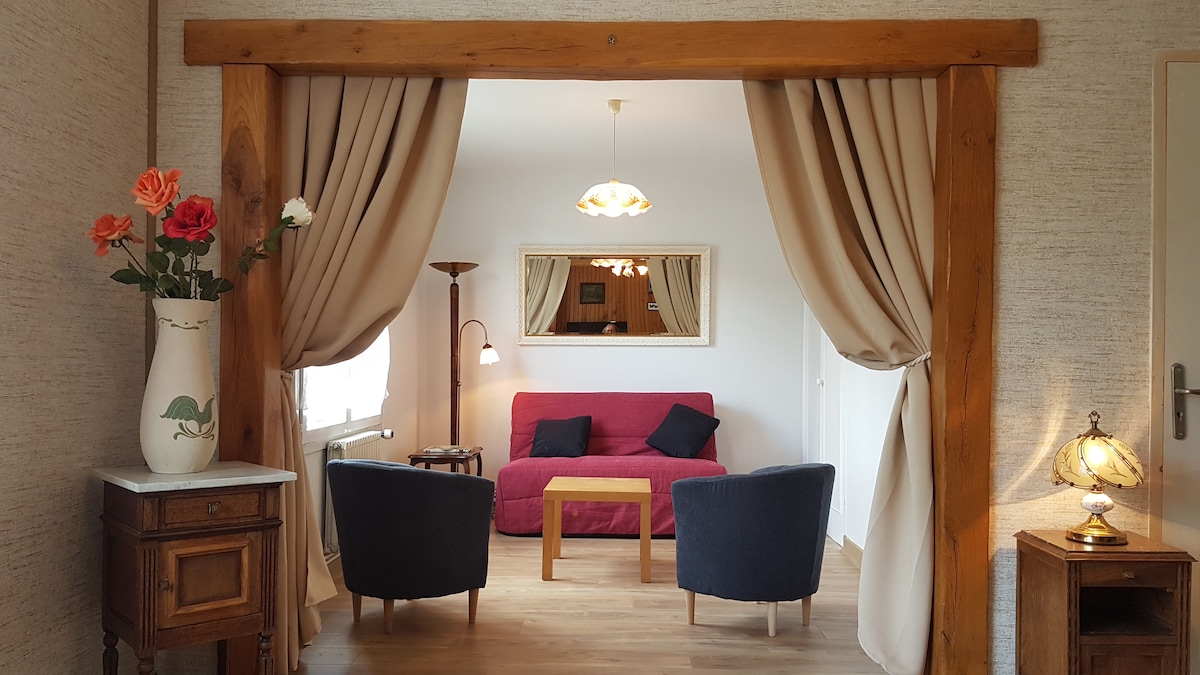 The living area is framed by wooden beams and neutral curtains, creating an inviting transition. A comfortable red sofa is paired with two dark armchairs and a wooden coffee table. A decorative lamp and a wall-mounted mirror enhance the natural light coming from the window.