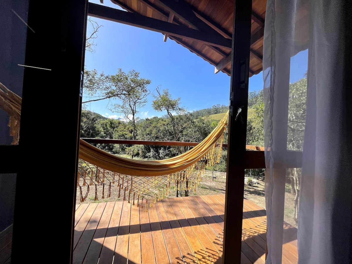 A private balcony features a vibrant yellow hammock suspended between wooden support beams. The wooden floorboards are visible, along with a stunning view of lush greenery and trees extending into the distance under a clear blue sky.