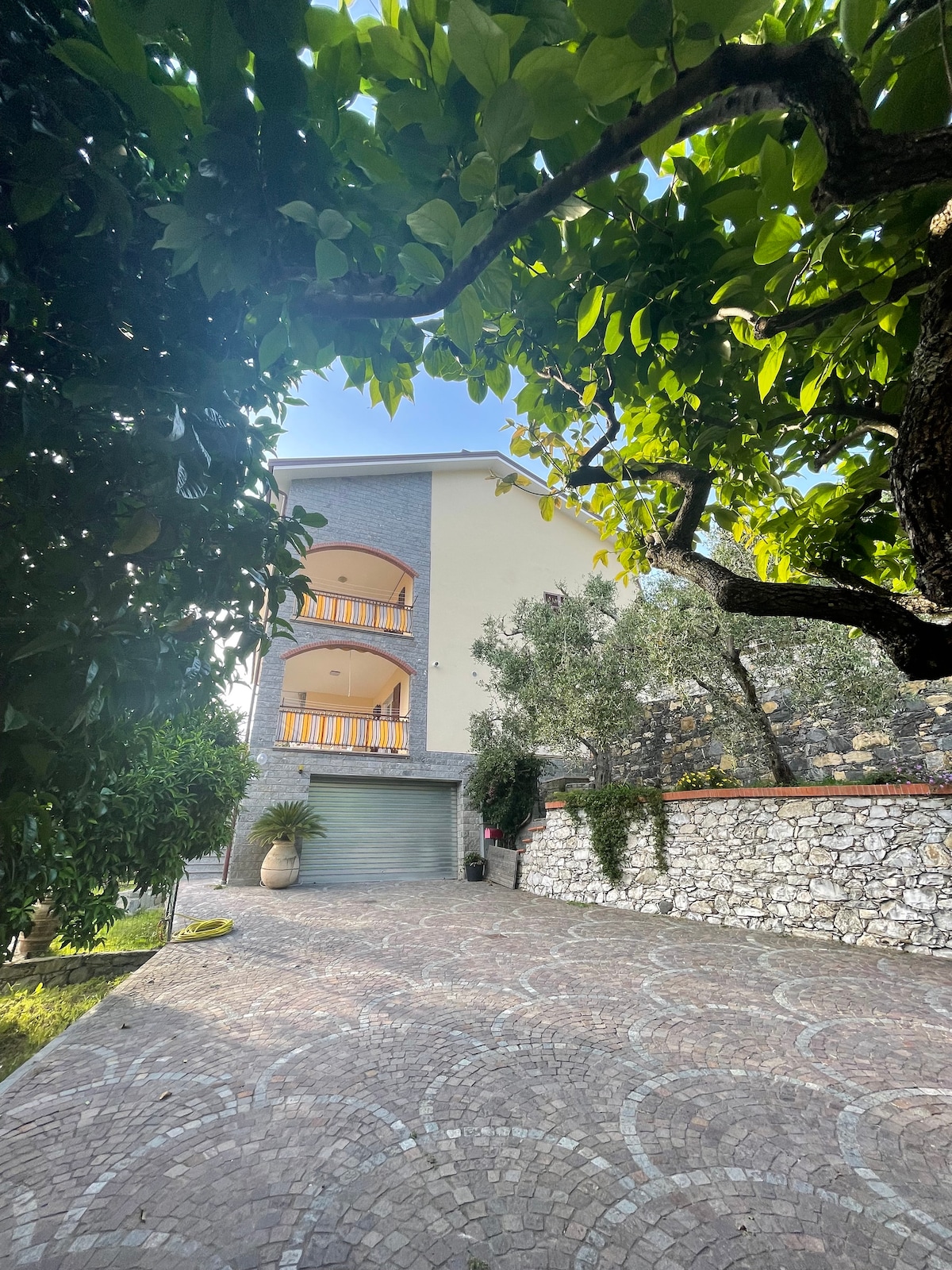 The exterior of a three-story house is framed by lush greenery, showcasing arched balconies on the upper floor. The driveway, made of patterned stones, leads up to a garage, while vibrant foliage creates a natural canopy over the entrance, enhancing the tranquil setting.